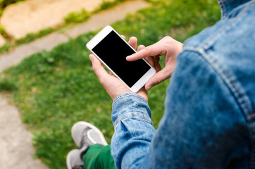 Man use his Mobile Phone outdoor, close up. Mobile phone in hands a young hipster business man in denim shirt and green jeans on the background of green grass.