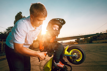 Trainer teaches small bikers to do stunts on a motorcycle. Small boy dressed in protective suit and helmet.