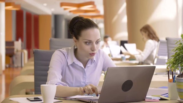 young happy woman shouts and laughs when see good news of success in computer during working day with papers sitting at table in co working colorful business office space