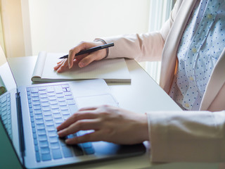 asian business woman(30s to 40s) record on blank book by black pen with pink or pastel suits with soft focus left hand on keyboard of laptop foreground