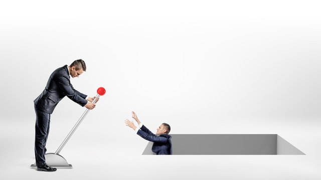 A businessman turns a large lever helping another man fallen inside a large square hole in the ground.