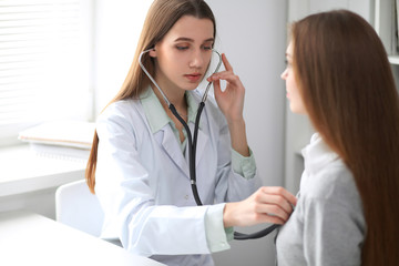 Female doctor examining her patient with stethoscope while sitting at the table near the window in hospital. Physician is ready to help patient. Medicine and health care concept