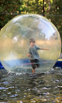 Five year old boy in shorts and grey t shirt balancing inside a huge zorb ball on the water in the summer amusement park. Water walking or zorbing in the theme park