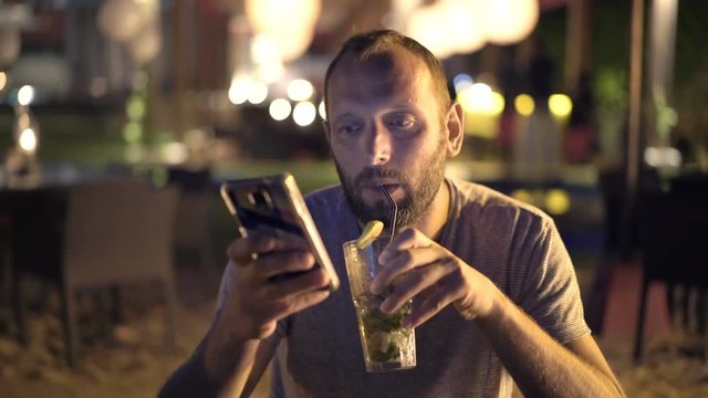 Young man drinking cocktail and texting on smartphone in outdoor bar at night 
