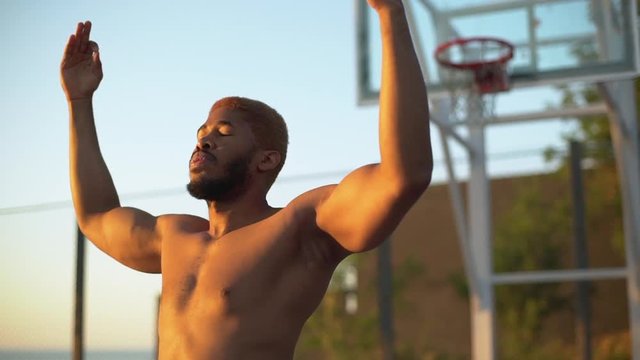 slow motion in early morning young  african american man with naked body putting hands up and meditating on basketball court under sunshine