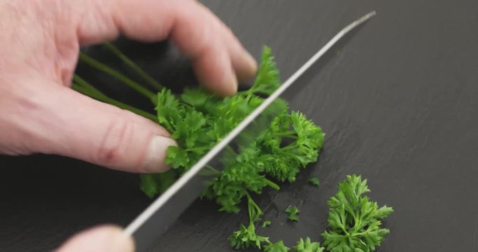 A man cutting curly parsley on a black slate stone cutting board with a large old knife.