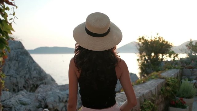 Brunette with luxurious curls and hat stand with back and look at sea.