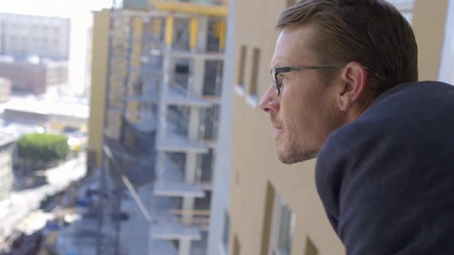 Developer, architect or business man in a suit leans out of window and looks at cityscape including nearby construction. Medium close up profile, hand-held real-time 4K.