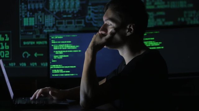 portrait of tired young programmer working at a computer in the data center filled with display screens