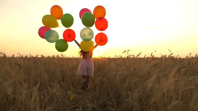 young girl in the dress with colorful ballons is running across the field.