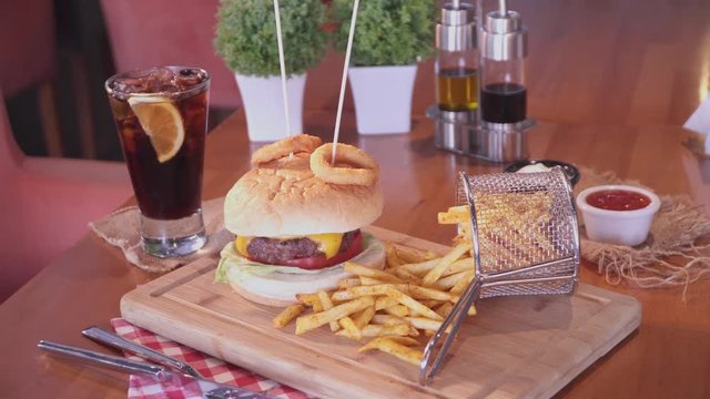 American cheeseburger with cheese, beef, tomato, lettuce, onion rings with sticks, french fries and cold drink soda with lemon slice. Pan shot.