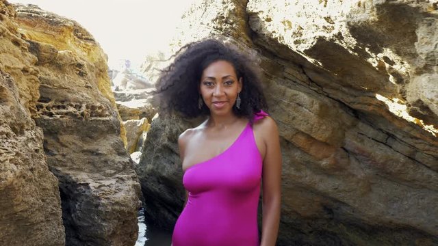 Portrait of black people, pretty happy young african american woman looking at camera and smiling in canyon..Rocks beach.Sexy mixed race girl in pink swimsuit and jewelry enjoying nature. 4k
