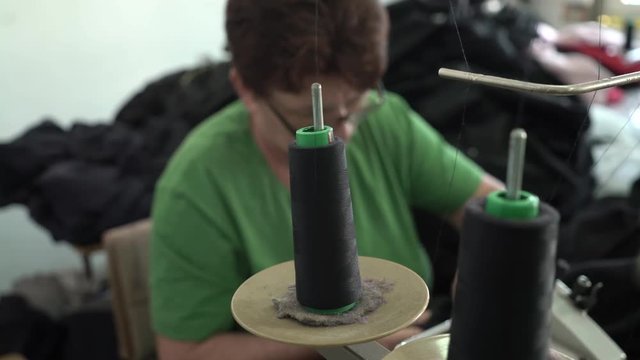 Sewing machine pulls threads from the spools in focus, mature Caucasian woman tailoring in the blurred background, steadicam shot from the female tailor, close up, room interior