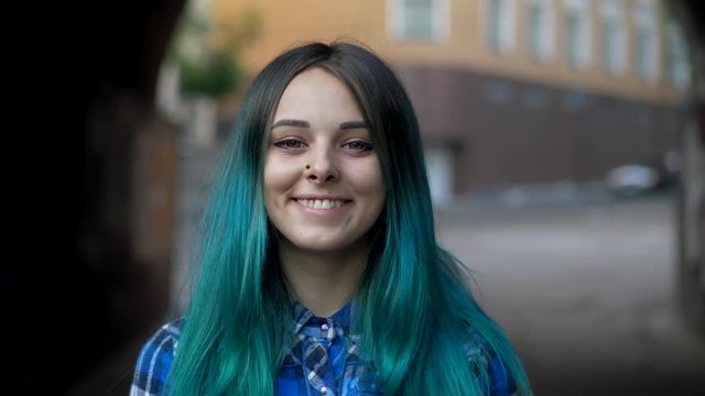 Street trendy or hipster girl with blue dyed hair. Woman with piercing in nose, violet lenses, ears tunnels and unusual hairstyle stands in city. 4k
