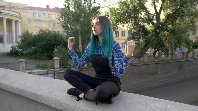 Calm street hipster girl with blue dyed hair. Woman with piercing in nose, unusual hairstyle meditating on European empty street. Yoga concept. 4k