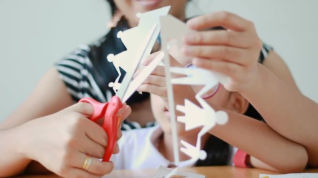 Asian little girl in Thai kindergarten student uniform and her mother using scissor to cut the white paper making family shape father mother son and daughter on wooden table select focus on hands
