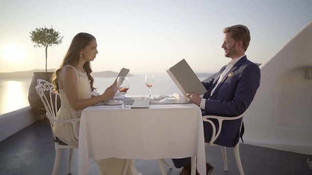 4k travel video dolly move beautiful honeymoon couple at dinner table on terrace above sea outdoors of restaurant in santorini island during romantic sunset