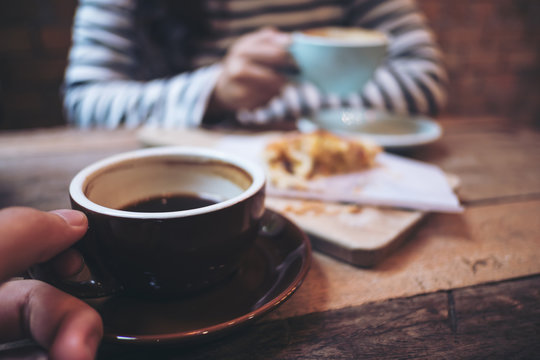 Closeup image of a man and a woman holding and drinking hot coffee together with a piece of raisin danish on wooden vintage table in coffee shop