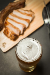 Beer in a tall glass and delicious sliced pork chop steak with spices and grilled garlic on wooden board (selective focus); top view on black table background.