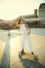 girl in a white dress and with a longboard on the sunny square of the old city