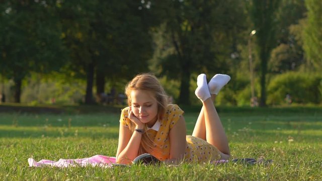 delayed shooting. A young blond girl with European wreath lies on her stomach in a clearing. on a warm sunny day in a meadow in the park. read book. a woman relaxes in the rays of the setting sun and