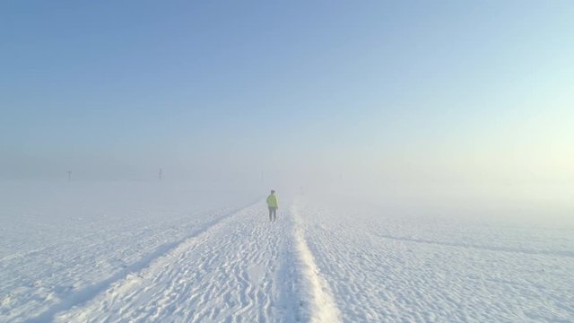 AERIAL: Unrecognizable woman walking along the snowy path through misty fields on beautiful winter day. Senior female walking across countryside covered in snow in foggy winter