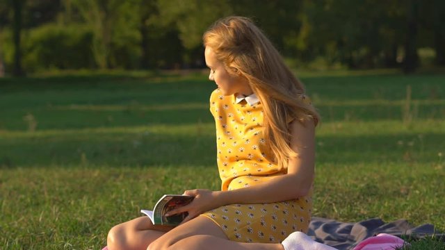 Slow motion. A young blonde girl in a warm sunny day at a picnic. The girl is dressed in a bright yellow dress in flowers. green grass . holds in his hand a book and reads. rest in the park. HD