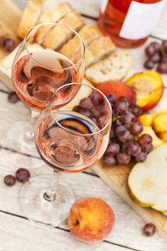 Two glasses of rose wine and board with fruits, bread and cheese on wooden table, shallow DOF