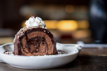 A piece of chocolate roll cake and whipped cream on the top in white ceramics plate and coffee cup on wooden table with blur background in cafe