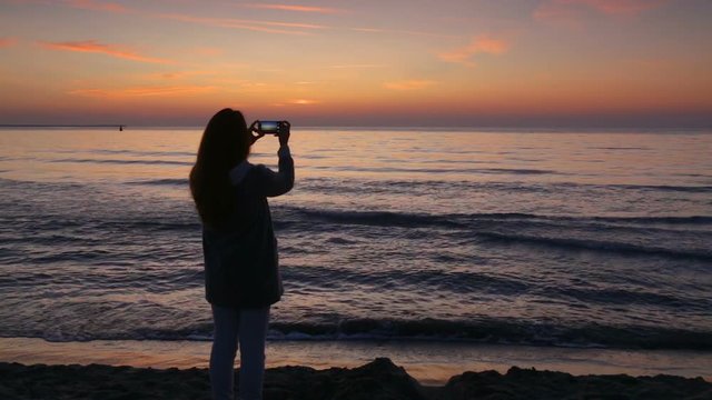 Young woman taking photos with her smartphone, on the beach, at sunrise