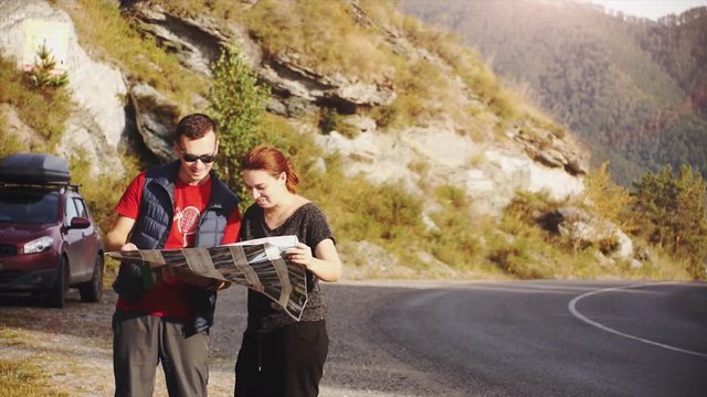 Tourists are guided by the terrain with a map. Young beautiful couple stand in amazing picturesque place amidst the mountains next to the car looking for right the way. 3840x2160
