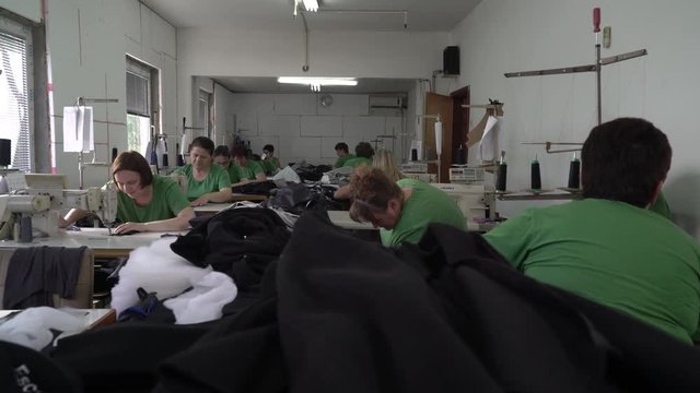 Group of female dressmakers sitting and using sewing machines to stitching clothes in workshop, wide angle view, young and mature women tailoring in clothing factory, interior scene, steadicam shot