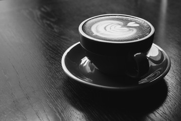 A cup of hot latte art or cappuccino coffee on wooden table in cafe. Black and white photo