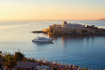 Casino building and white yacht on calm sea level in golden morning sun. St. Georges ´s bay, St. Julian ´s, Malta.