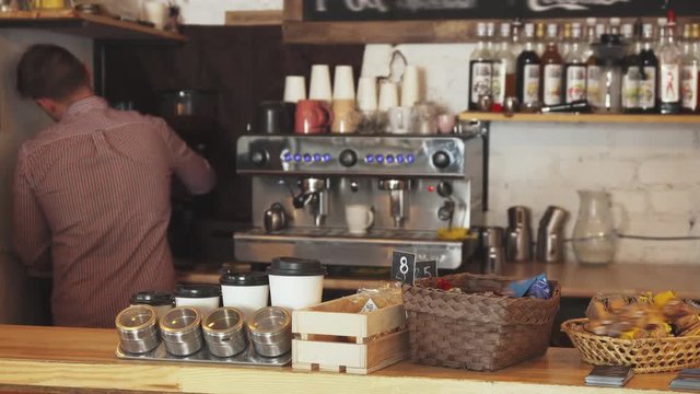 The barista is standing at the bar. He is making coffee. On the bar counter lie various spices and desserts. The barista works in the cafe