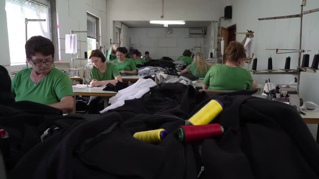 Colorful spool threads on the black fabric textiles in the foreground, group of female dressmakers stitching at sewing machines in workshop, steady cam shot, camera motion to the women, room interior