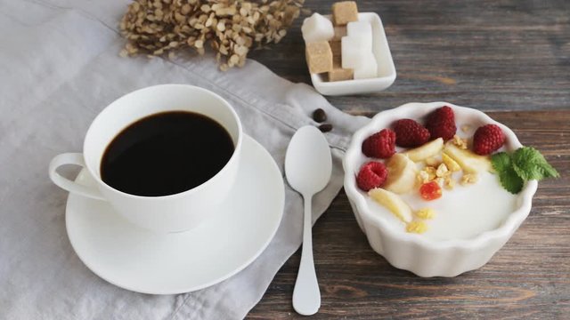 Concept of healthy organic eating - breakfast on wooden table - cup of coffee, yogurt with cereals, raspberry berries and sugar cubes. Camera locked down.