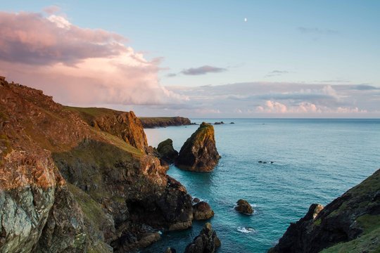 A view towards Lands End from Kynance Cove in Cornwall, UK showing harsh rugged rocks and cliffs at sunset with calm and deep blue ocean to the horizon.