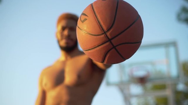 closeup slow motion in summer time handsome bearded man with naked torso holding basketball and showing it to camera standing on court