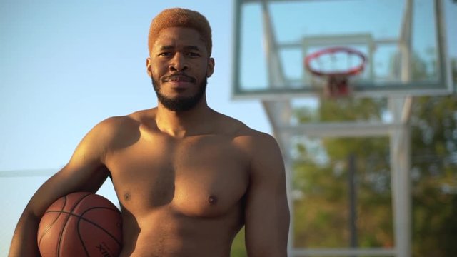 closeup slow motion at sunrise muscular guy with dyed hair and naked torso looking at camera smiling standing on court holding  basketball background in defocus 