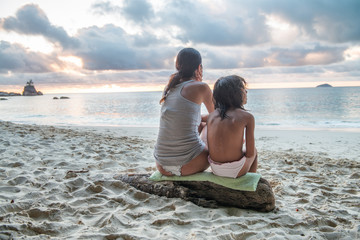 Mother and daughter relaxing on the beach at sunset, back view