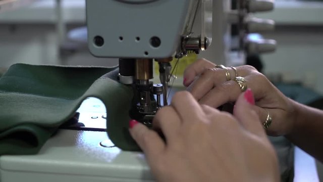 Woman hands close up during works at sewing machine, dressmaker stitches green factory textiles, shallow depth of field,concept tailoring, room interior