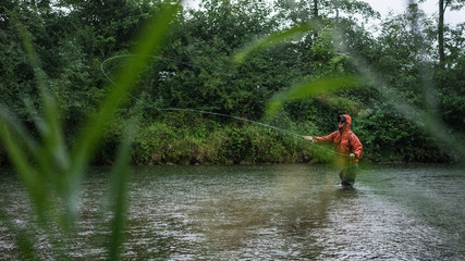 Fototapeta na wymiar Angler mit Wathose und Watjacke im Wasser beim Angeln mit Fliegenrute bei Regen im klaren Fluss stehend