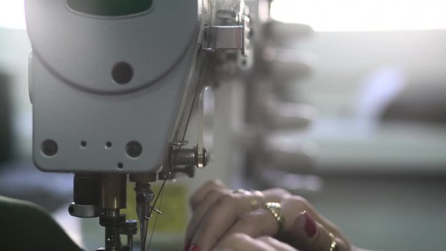 Female tailor works at sewing machine tilt down, hands of dressmaker stitches green cloth, shallow depth of field,concept tailoring, room interior, close up