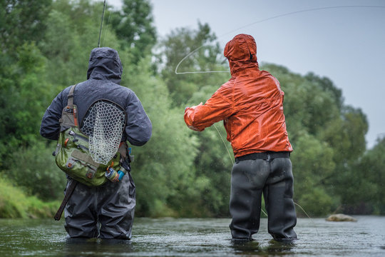 Angler mit Wathose und Watjacke im Wasser beim Angeln mit Fliegenrute bei Regen im klaren Fluss stehend und werfend