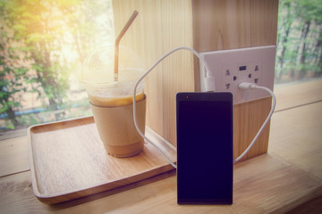 Smart phone while charging battery with ice coffee and natural background.(Vintage and Dream tone)