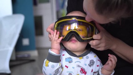 Smiling mother puts on ski goggles on baby with surprised face and eyes wide opened. Handheld shot