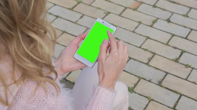 Young Woman in pink jeans sitting on bench uses SmartPhone with pre-keyed green screen. Few types of gestures - scrolling up and down, tapping, zoom in and out. Perfect for screen compositing