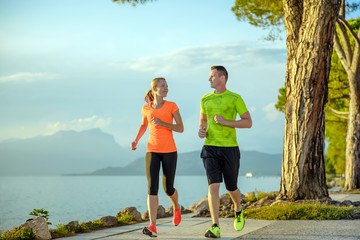 Young sexy couple is running along the promenade. they are doing their sport workout in the beautiful sundown along the beach. colorful dress, trees, water, mountains and a amazing blue sky.
