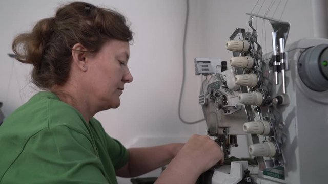 Portrait of middle aged Caucasian woman during sits at sewing machine and tailoring, dressmaker stitching cloth in workshop, face close up, interior scene